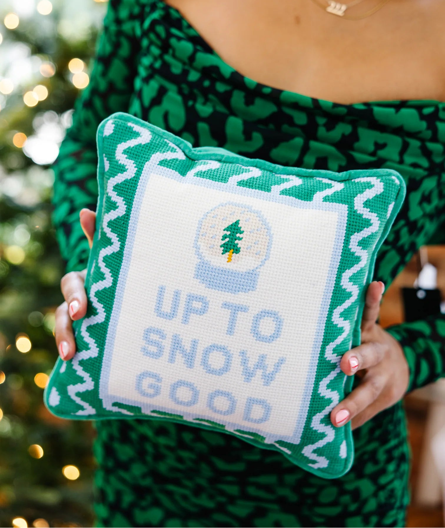Person holding a green pillow with 'Up to Snow Good' text against a blurred Christmas tree background